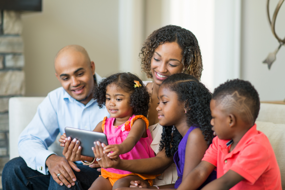 A smiling family of five sits together on a couch, looking at a tablet. The mother and father are seated behind their three young children, who are engaged and smiling as they hold the device. The cozy, well-lit living room setting conveys warmth, connection, and shared enjoyment of technology.