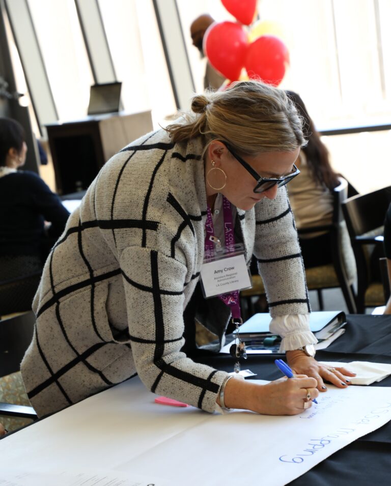 Amy Crow, Assistant Regional Administration for LA County Library, leans over a table, writing "Asset Mapping" on a large white paper with a blue marker at an event.