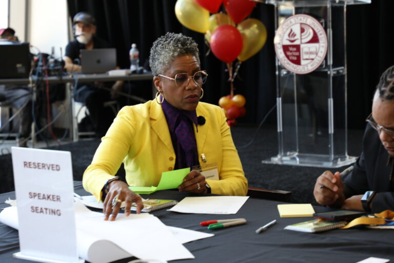 A woman in a yellow blazer and glasses sits at a table, holding a green paper. A 'Reserved Speaker Seating' sign is beside her. In the background, a podium with the CSU Dominguez Hills logo and balloons are visible.