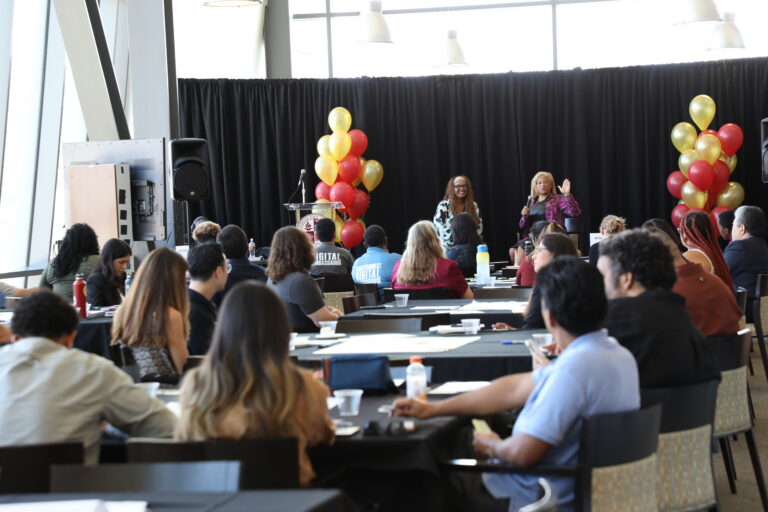 Two women present on stage to a diverse audience seated at tables. One speaker holds a microphone and gestures, while the other stands beside her. Red and gold balloons decorate the stage.