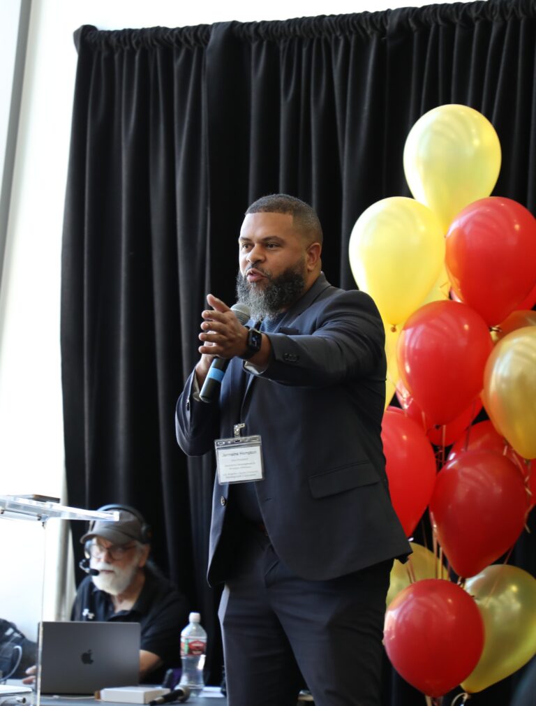 a man with a beard wearing a dark suit, speaks into a microphone with his hand gesturing. Red and yellow balloons are to his right, and black curtains are in the background.
