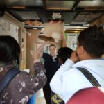 Instructor demonstrating roofing materials to a small group inside a training space, holding up samples while participants observe and ask questions.