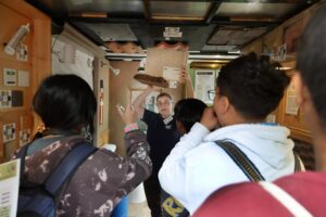 Instructor demonstrating roofing materials to a small group inside a training space, holding up samples while participants observe and ask questions.